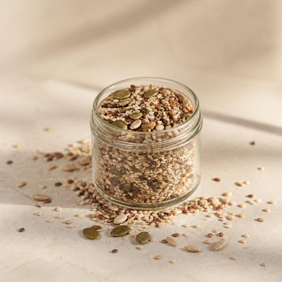 Mixed seeds and grains in a small glass jar on a light sand surface